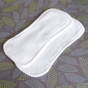 Peut inclure: Deux coussinets rectangulaires blancs aux bords arrondis sont empilés. Ils sont faits d'un matériau doux et absorbant. Le fond est un tissu gris avec un motif répété de feuilles vertes et blanches.