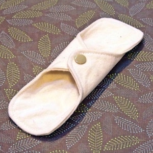May include: A white cloth menstrual pad with a snap closure. The pad is folded in half and is lying on a patterned fabric background.