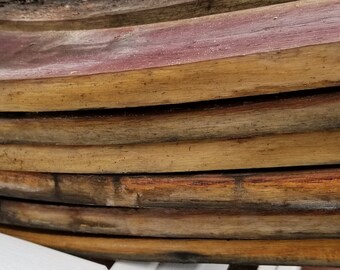 May include: Close-up of a stack of wooden wine barrel staves. The staves are a light brown color with some darker brown streaks and spots. The staves are curved and have a rough texture.