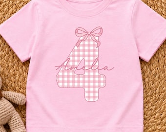 May include: A light pink short-sleeved t-shirt featuring a large number four with a white and pink gingham pattern. The number is topped with a pink line-art bow and the name Amelia is written in a cursive script across the center of the number.