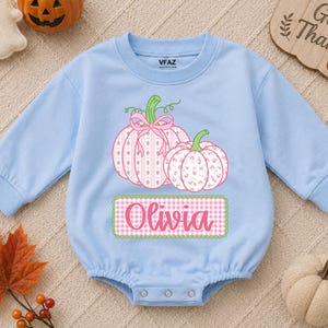 May include: A light blue long-sleeved baby romper featuring a graphic of two pumpkins with pink floral and plaid patterns, a pink bow, and a green-bordered rectangular label with the name Olivia in pink script text on the front.