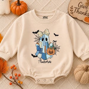 May include: A beige long-sleeve baby bodysuit featuring a graphic of a blue patchwork ghost wearing a bow, holding a jack-o'-lantern, and surrounded by spiderwebs and bats. The name Aurora is printed below the ghost in a black script font.
