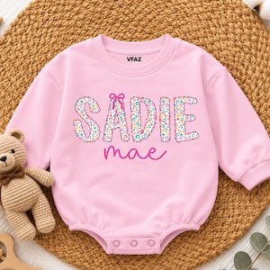 May include: A light pink long-sleeve baby bodysuit featuring the name SADIE in colorful leopard print letters with a small pink bow above the letter I, and the name mae in cursive pink script below. The brand name VFAZ is printed at the neck.