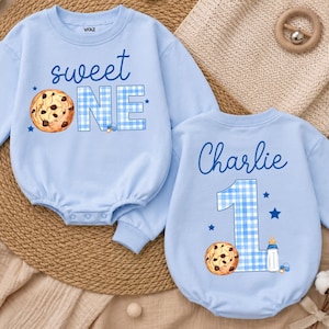 May include: Two light blue long-sleeve baby rompers. One features the text "sweet ONE" with a chocolate chip cookie and blue gingham pattern. The other features the name "Charlie" above a large blue gingham "1", a cookie, and a baby bottle.
