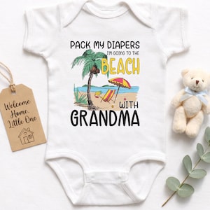 May include: White baby onesie with a beach-themed graphic. The design includes a palm tree, beach umbrella, and the text "PACK MY DIAPERS I'M GOING TO THE BEACH WITH GRANDMA." A small teddy bear and a tag that says "Welcome Home, Little One" are also present.