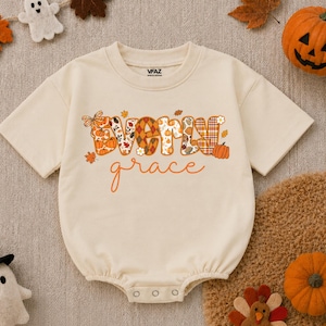 May include: A cream-colored short-sleeve baby romper featuring the text Everly grace in a decorative autumn-themed font. The letters are filled with patterns of pumpkins, plaid, and fall leaves. The romper has a round neckline and snap closures.