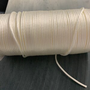 May include: A roll of off-white cord, tightly wound and showing a smooth texture with a subtle sheen. The cord is a neutral colour, suitable for various crafting projects. The roll is on a dark, textured surface.