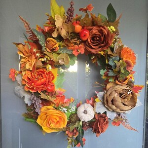 Könnte beinhalten: Ein runder Kranz mit künstlichem Herbstlaub, bestehend aus orangefarbenen und gelben Rosen, dunkelroten Pfingstrosen, kleinen roten Äpfeln, einem weißen Miniaturkürbis, grünen Blättern und braunen Farnakzenten in einer dichten Anordnung.