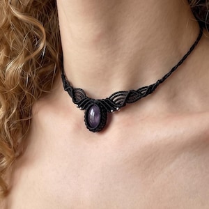 May include: A black macrame choker necklace featuring a central oval-shaped purple gemstone. The cord is intricately knotted in a symmetrical pattern around the stone, extending into a thin, braided band that secures around the neck.