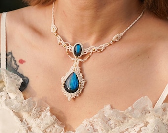 May include: A handcrafted macrame necklace featuring a teardrop-shaped blue gemstone pendant and a smaller oval gemstone. The necklace is made with cream-colored cord and gold-colored accents, creating an elegant and bohemian style.