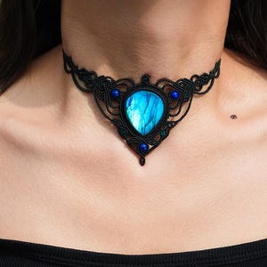 May include: A black macrame choker necklace featuring a large, oval, blue gemstone pendant. The pendant is surrounded by intricate macrame detailing and accented with smaller blue beads. The necklace is worn around the neck.