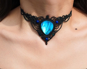 Puede incluir: Un collar gargantilla de macramé negro con un colgante grande, ovalado, de piedra preciosa azul. El colgante está rodeado de intrincados detalles de macramé y acentuado con cuentas azules más pequeñas. El collar se lleva alrededor del cuello.