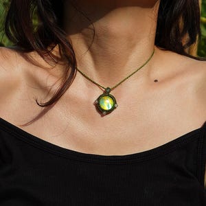 May include: A handmade macrame necklace with a round pendant. The pendant features a central, iridescent stone with shades of yellow and green, surrounded by a woven, olive-green macrame setting. The necklace is worn on a black tank top.