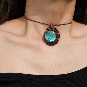 May include: A brown macrame choker necklace with a round, iridescent blue-green gemstone pendant. The pendant is set within a crescent-shaped macrame frame. The necklace is worn around the neck.