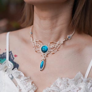 May include: A cream macrame necklace with a large, round, blue gemstone centre and a teardrop-shaped pendant. The necklace features intricate knotting and small gold-coloured beads. The necklace is worn on a person wearing a white top.