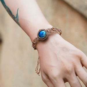 May include: A macramé bracelet with a large, oval, blue gemstone at its centre. The bracelet is made of tan-coloured woven cord with small gold-coloured beads. The gemstone has a shimmering, iridescent quality. The bracelet is worn on a wrist.