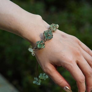 May include: A handmade bracelet featuring green gemstones encased in a woven, brown cord. The bracelet has a natural, earthy aesthetic, with several larger stones and smaller, dangling accents. The design is simple yet elegant, suitable for everyday wear.