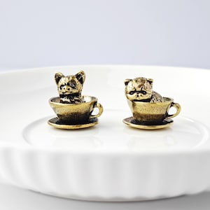May include: Two small, antique gold-colored cat figurines sit inside teacups on saucers. The cats are detailed with textured fur and facial features. The teacups have handles and sit on a white, fluted plate.