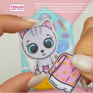 Op de afbeelding: Een grijze en witte kattenpoppenhuis papieren uitsnede met grote blauwe ogen en een roze luier. De poppenhuis uitsnede wordt vastgehouden door een hand. De poppenhuis uitsnede maakt deel uit van een grotere set poppenhuis uitsnedes. De tekst "YELLOW DUCK DOLLS" is zichtbaar in de linkerbovenhoek van de afbeelding.