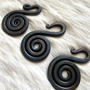 May include: Three handcrafted, dark metal spiral hooks with a rustic finish. Each hook features a circular spiral design with a curved hook end. The hooks are displayed on a white, textured surface, showcasing their unique, artisan craftsmanship.