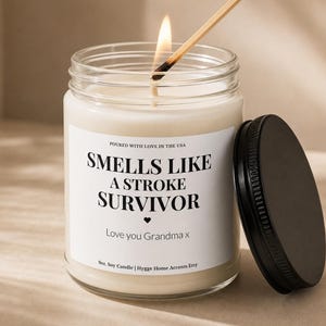May include: A lit soy candle in a glass jar with a black lid. The candle has a white label with the text "Smells Like A Stroke Survivor" and "Love you Grandma x". A match is being used to light the candle.