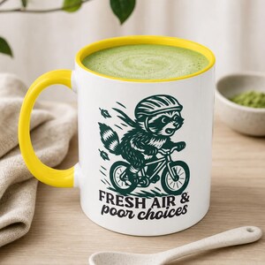 May include: White ceramic mug with a yellow handle and rim, filled with green liquid. The mug features a raccoon riding a bicycle with the text "FRESH AIR & POOR CHOICES". A white spoon rests on a wooden surface.