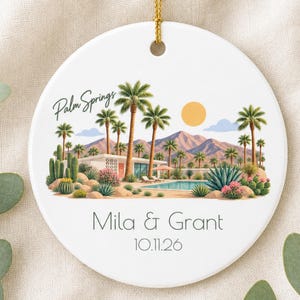 May include: A round white ceramic ornament with a gold string featuring a colorful illustration of a Palm Springs desert landscape with palm trees, a swimming pool, mountains, and a sun. The text reads Palm Springs, Mila & Grant, 10.11.26.