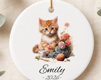 May include: A round white ceramic ornament hanging from a gold string. The center features an illustration of a fluffy orange kitten sitting next to a small cup, pink flowers, and a ball of yarn. The name Emily and the year 2026 are printed below.