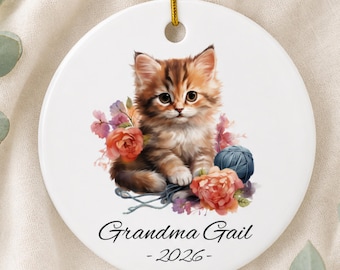 Pode incluir: Um ornamento redondo de cerâmica branca com um gatinho malhado castanho sentado entre flores cor-de-rosa e cor de laranja e um novelo de lã azul. O texto "Grandma Gail" e "2026" está impresso em letra preta por baixo do gatinho. Cordão dourado.