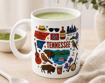 Puede incluir: Taza de cerámica blanca con una bebida verde en su interior. La taza está decorada con ilustraciones coloridas de símbolos de Tennessee, incluyendo una guitarra, un bisonte y la palabra "Tennessee". El diseño incluye un sombrero, gafas de sol y una botella de whisky.