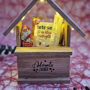 May include: A wooden Christmas advent calendar with a star on top and lights inside. The calendar is filled with small gifts, including a chocolate Santa Claus, a box of tea, and a tube of hand cream. The calendar is decorated with the words "Adventszauber" which means "Advent Magic" in German.
