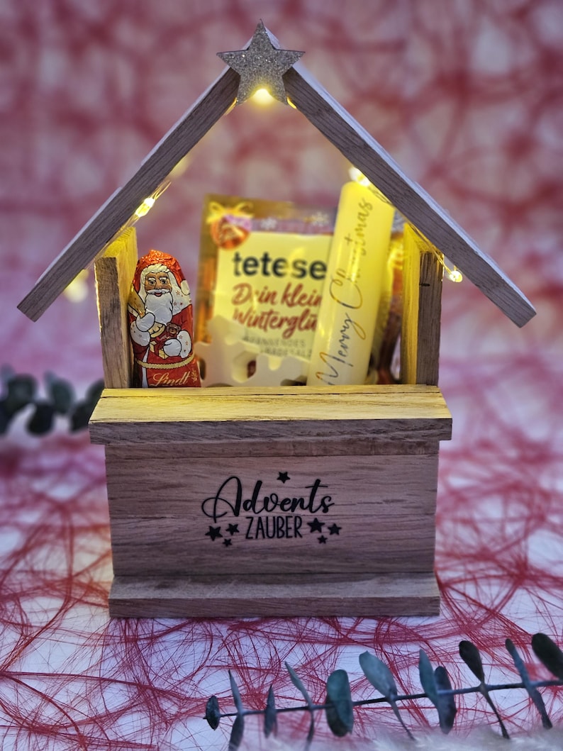 May include: A wooden Christmas advent calendar with a star on top and lights inside. The calendar is filled with small gifts, including a chocolate Santa Claus, a box of tea, and a tube of hand cream. The calendar is decorated with the words "Adventszauber" which means "Advent Magic" in German.