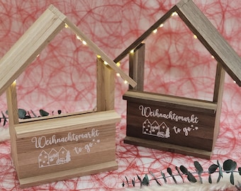 May include: Two wooden Christmas market-themed decorative houses with string lights. One is light-colored wood, the other is dark. Each has the text "Weihnachtsmarkt to go" and a house graphic.