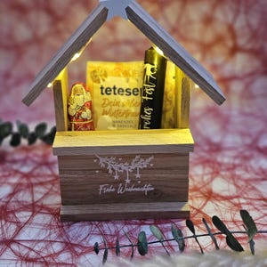 May include: A wooden Christmas village scene with a lit-up star on top. Inside the village is a small Santa Claus figurine, a box of tea, and a candle. The base of the village says "Frohe Weihnachten" in white lettering.