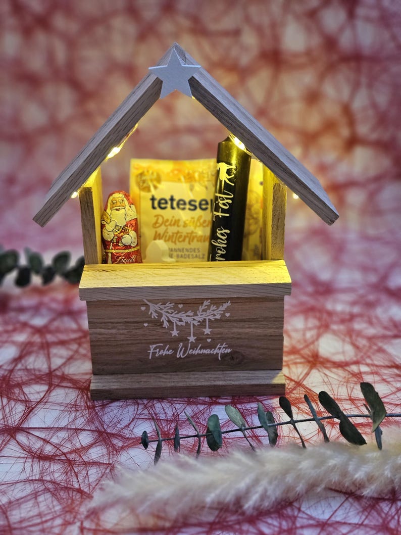 May include: A wooden Christmas village scene with a lit-up star on top. Inside the village is a small Santa Claus figurine, a box of tea, and a candle. The base of the village says "Frohe Weihnachten" in white lettering.
