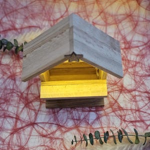 May include: A small wooden manger with a star on the roof and a light inside. The manger is sitting on a red and white textured surface.