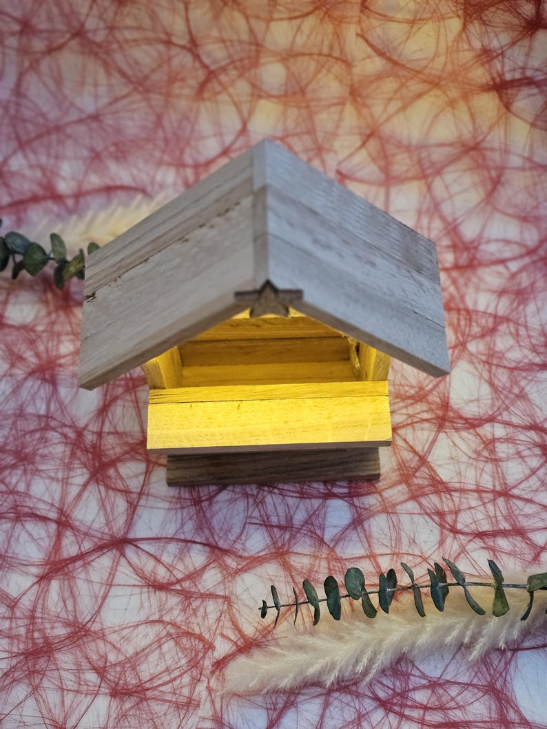 May include: A small wooden manger with a star on the roof and a light inside. The manger is sitting on a red and white textured surface.