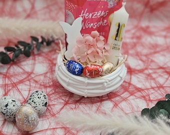 May include: A white ceramic nest with a white bunny figurine, pink flowers, chocolate eggs, and a white candle with a gold number one on it. The nest is sitting on a red and white shredded paper background.