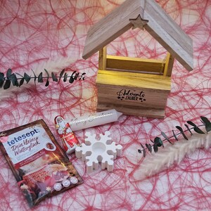 May include: A white wooden Christmas house with a lit candle inside and the text "Adventszauber" on the front. There is a white snowflake, a small Santa Claus chocolate, and a packet of tea with the text "tetesept Dein kleines Wintergl&uuml;ck" on a red and white background.
