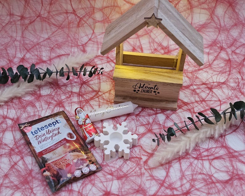 May include: A white wooden Christmas house with a lit candle inside and the text "Adventszauber" on the front. There is a white snowflake, a small Santa Claus chocolate, and a packet of tea with the text "tetesept Dein kleines Wintergl&uuml;ck" on a red and white background.