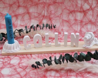 May include: White wooden name sign with the name "COLLIN" spelled out in white letters. A small blue candle with the text "WÜNSCH DIR WAS!" is in a white candle holder to the left of the name. The sign is on a red background.