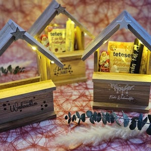 May include: Three wooden Christmas market stalls with lights, each decorated with a different festive design. The stalls are filled with small gifts and treats, including chocolate figures and a bag of hot chocolate mix. The text on the stalls reads "Frohe Weihnachten" and "Frohe Festtage".