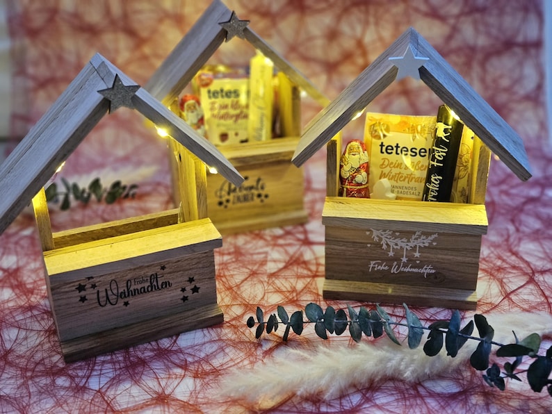 May include: Three wooden Christmas market stalls with lights, each decorated with a different festive design. The stalls are filled with small gifts and treats, including chocolate figures and a bag of hot chocolate mix. The text on the stalls reads "Frohe Weihnachten" and "Frohe Festtage".