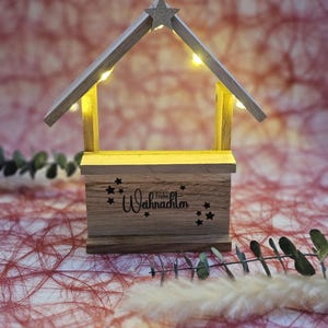 May include: A wooden Christmas manger with a star on top and lights inside. The manger has a sign that says "Frohe Weihnachten" which means "Merry Christmas" in German.