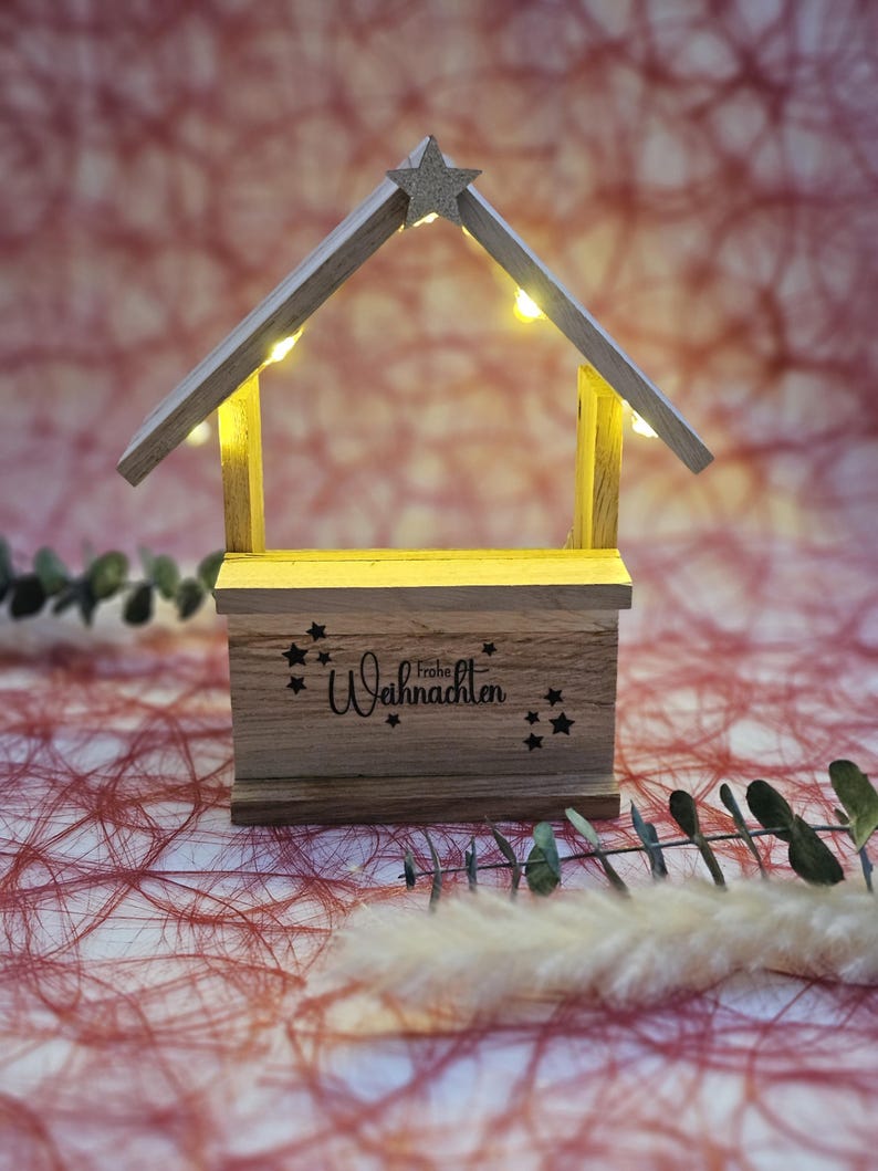 May include: A wooden Christmas manger with a star on top and lights inside. The manger has a sign that says "Frohe Weihnachten" which means "Merry Christmas" in German.