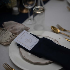 May include: A white plate with a gold rim and a navy blue napkin. The napkin is folded and has a white card on top of it. The card has the text "Passport" and a small airplane icon. The text "Matthew & Natalie" is also on the card.