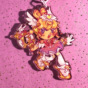 Op de afbeelding: Een heldere acryl bedel met een stripfiguur met blond haar, vleugels en een roze en witte outfit. Het personage houdt een goudkleurig en roze wapen vast. De achtergrond is een roze oppervlak met stervormige glitter.