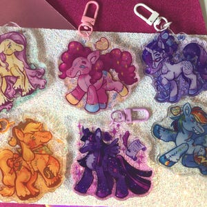 Puede incluir: Colección de seis llaveros acrílicos con temática de pony. Cada llavero presenta un diseño de pony diferente con colores vibrantes y cierres metálicos adjuntos. Los llaveros se muestran sobre una superficie brillante.