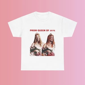 Puede incluir: Camiseta blanca con el texto "PROM QUEEN OF 1976" impreso en rojo. La camiseta presenta una foto de dos mujeres con vestidos de graduación y coronas, sosteniendo ramos de flores.