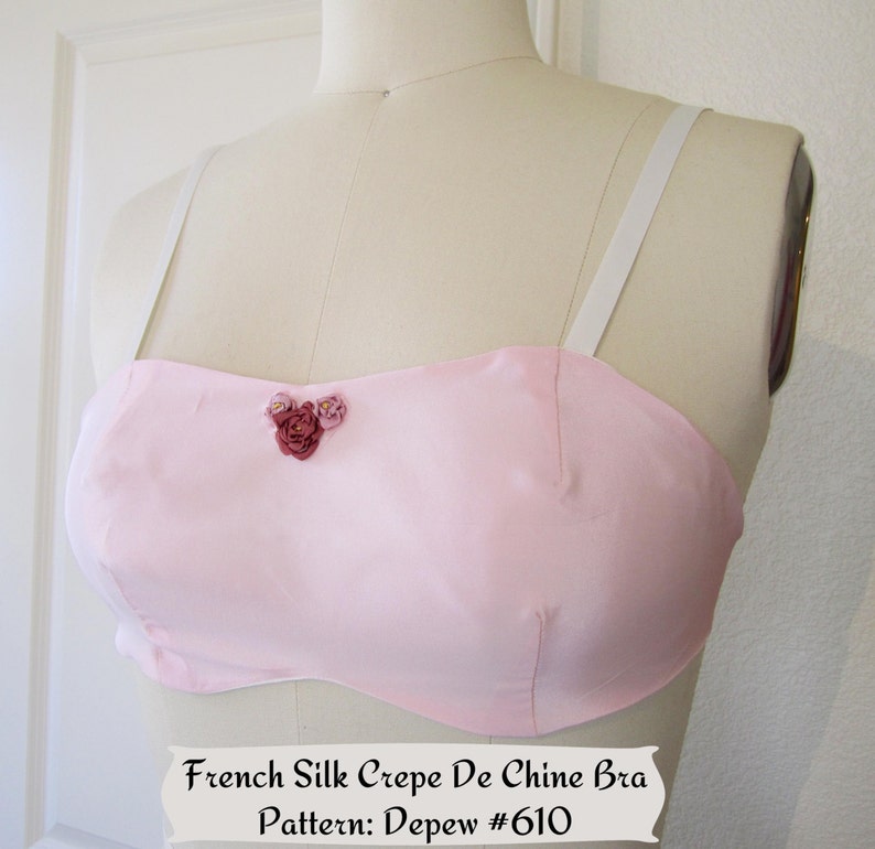 May include: A pink silk crepe de chine bra with white straps and a floral embellishment. French Silk Crepe De Chine Bra Pattern: Depew #610